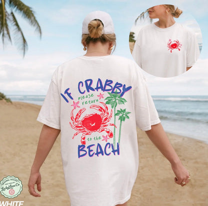 Person wearing a white t-shirt with a colorful crab design and text on a beach.