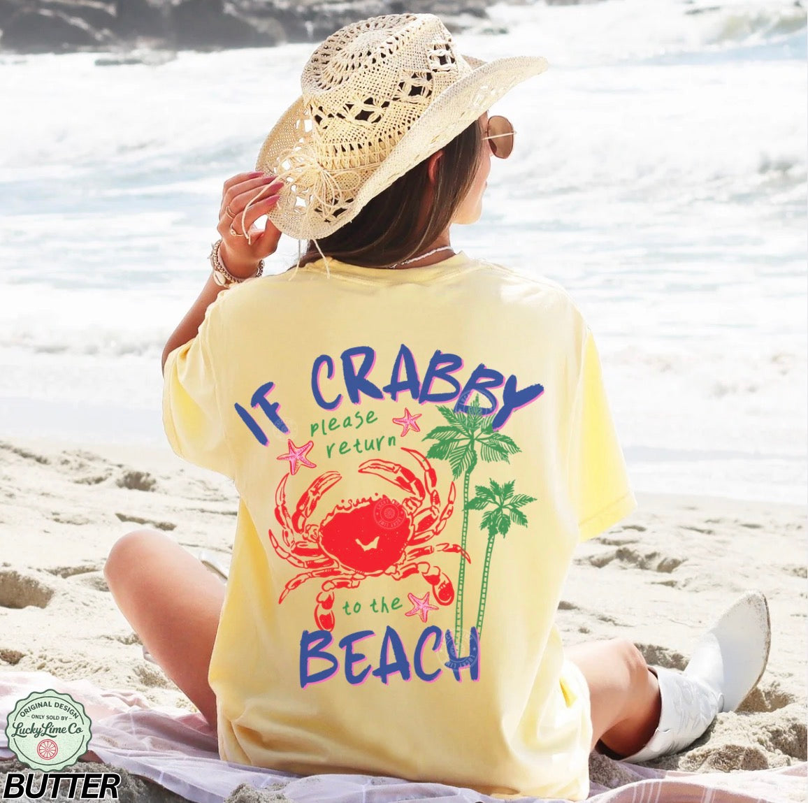 Person sitting on a beach wearing a yellow t-shirt with a crab and palm tree design.
