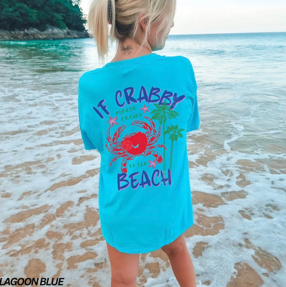 Person wearing a light blue t-shirt with a crab design and text on a beach.