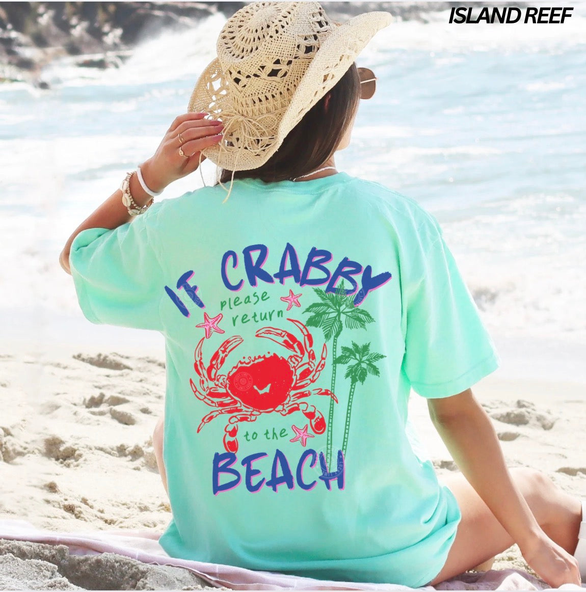 Person wearing a light blue t-shirt with a beach-themed design and 'Island Reef' branding on a beach.