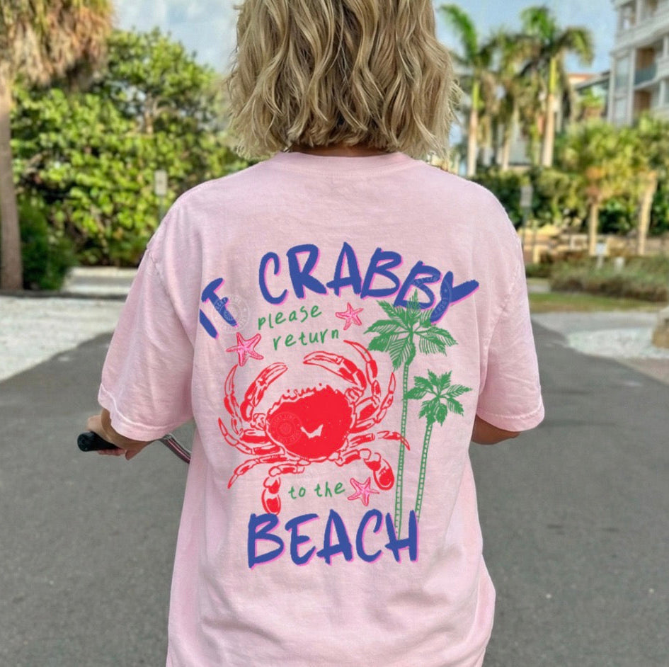 Person wearing a pink t-shirt with a beach-themed design and text, standing on a street.