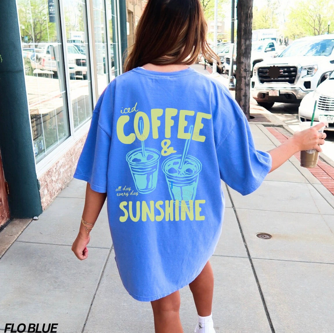 Person wearing a blue oversized t-shirt with coffee and sunshine design, standing on a sidewalk.