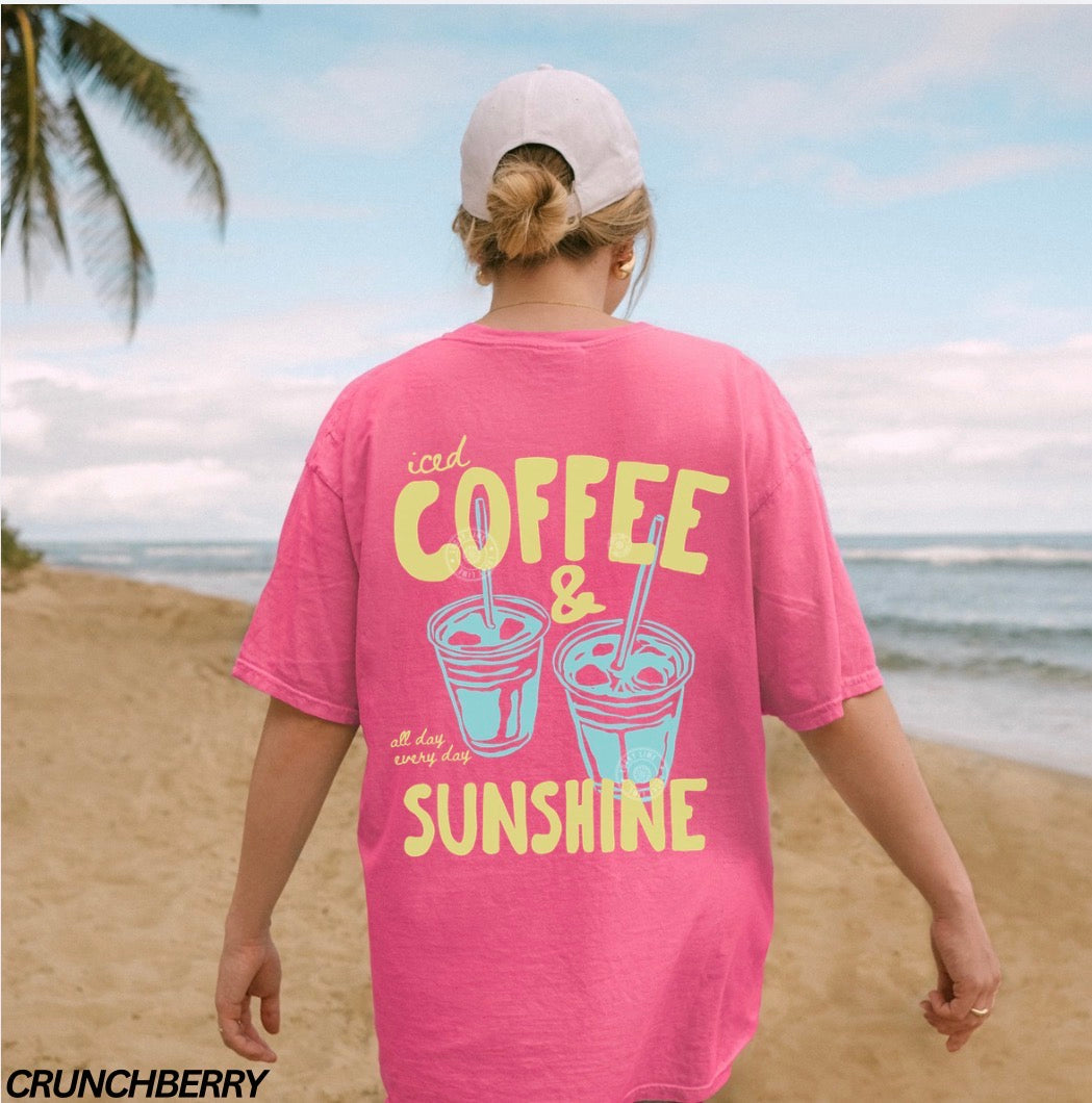 Person wearing a pink t-shirt with 'iced coffee & sunshine' design on a beach.