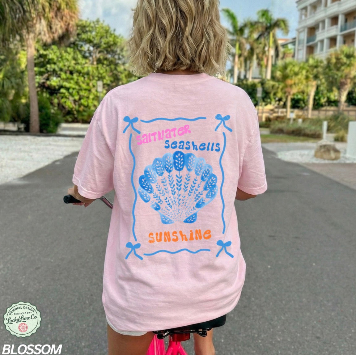 Person wearing a pink t-shirt with a shell design and text in an outdoor setting.