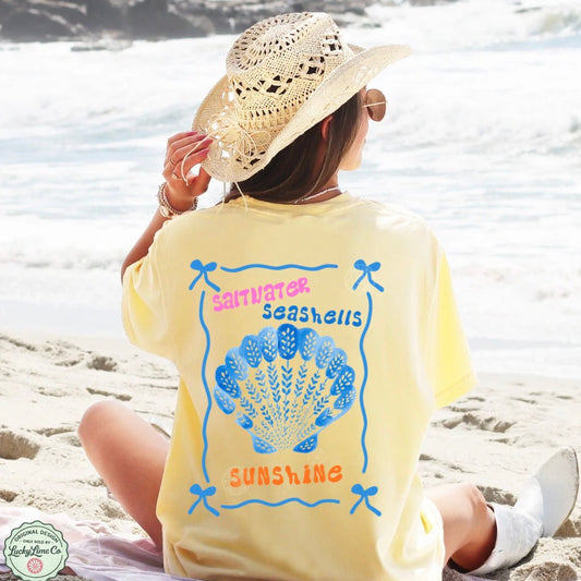 Person wearing a yellow t-shirt with a shell design and text on a beach.
