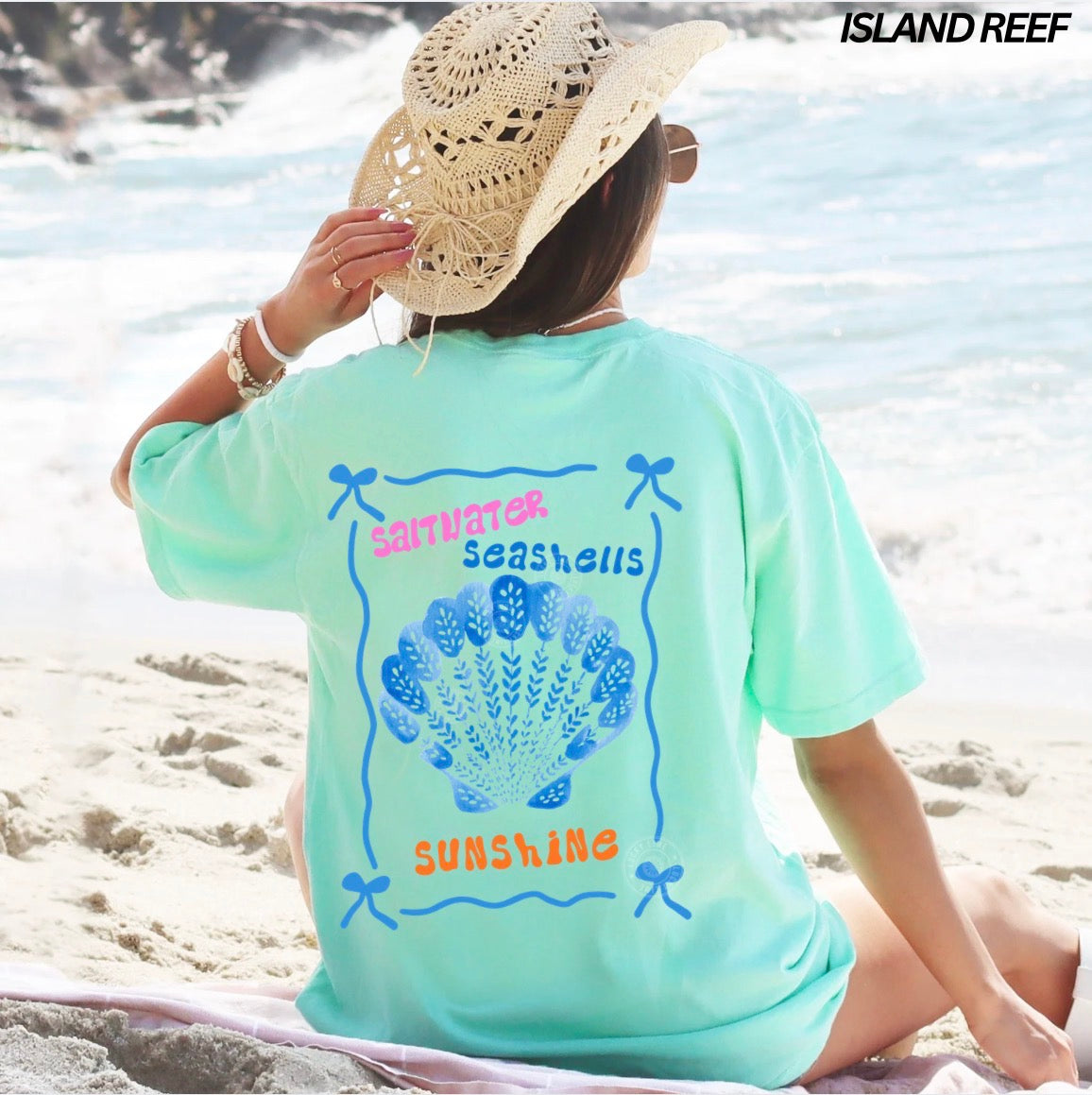 Person wearing a light blue t-shirt with a shell design and text on a beach, with 'Island Reef' branding.