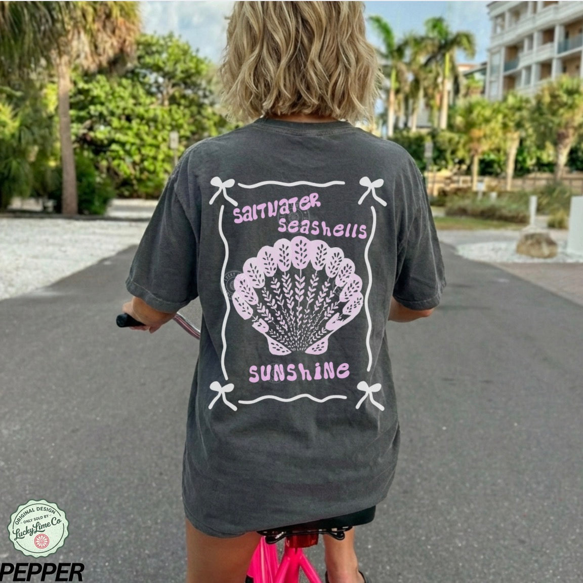 Person wearing a t-shirt with a shell design and text, riding a pink bike outdoors.