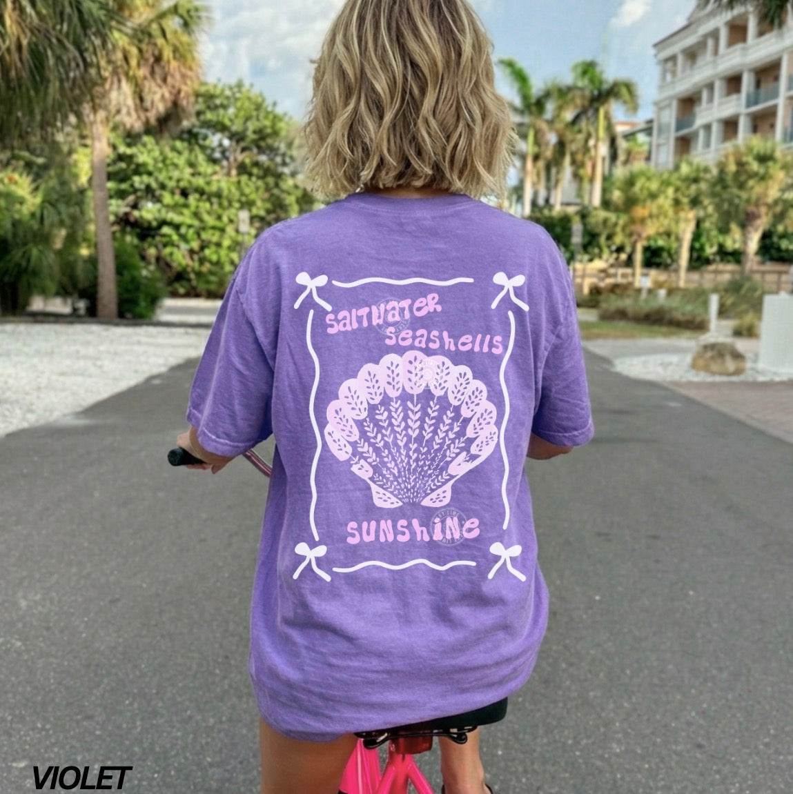 Person wearing a purple t-shirt with a white shell design and text, standing on a street.