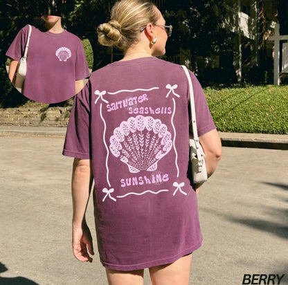 Person wearing a purple t-shirt with a shell design and text outdoors.