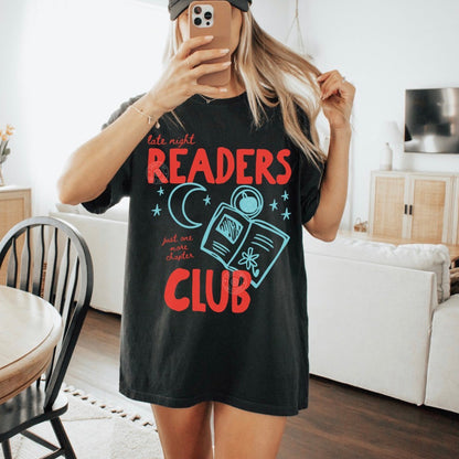 Person wearing a black t-shirt with 'late night readers club' design in a living room.