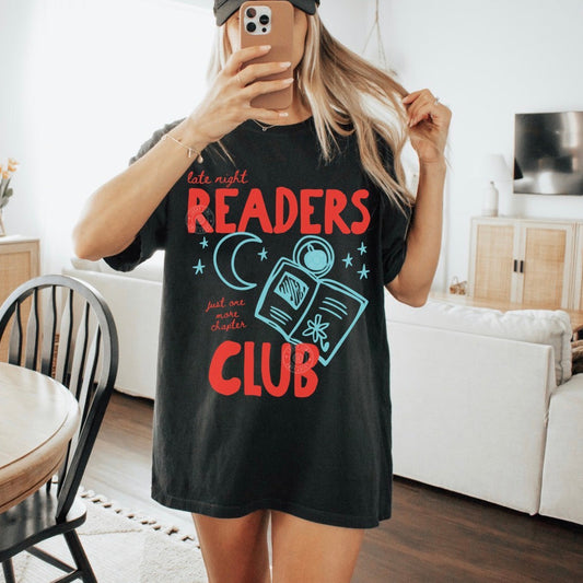Person wearing a black t-shirt with 'late night readers club' design in a living room.