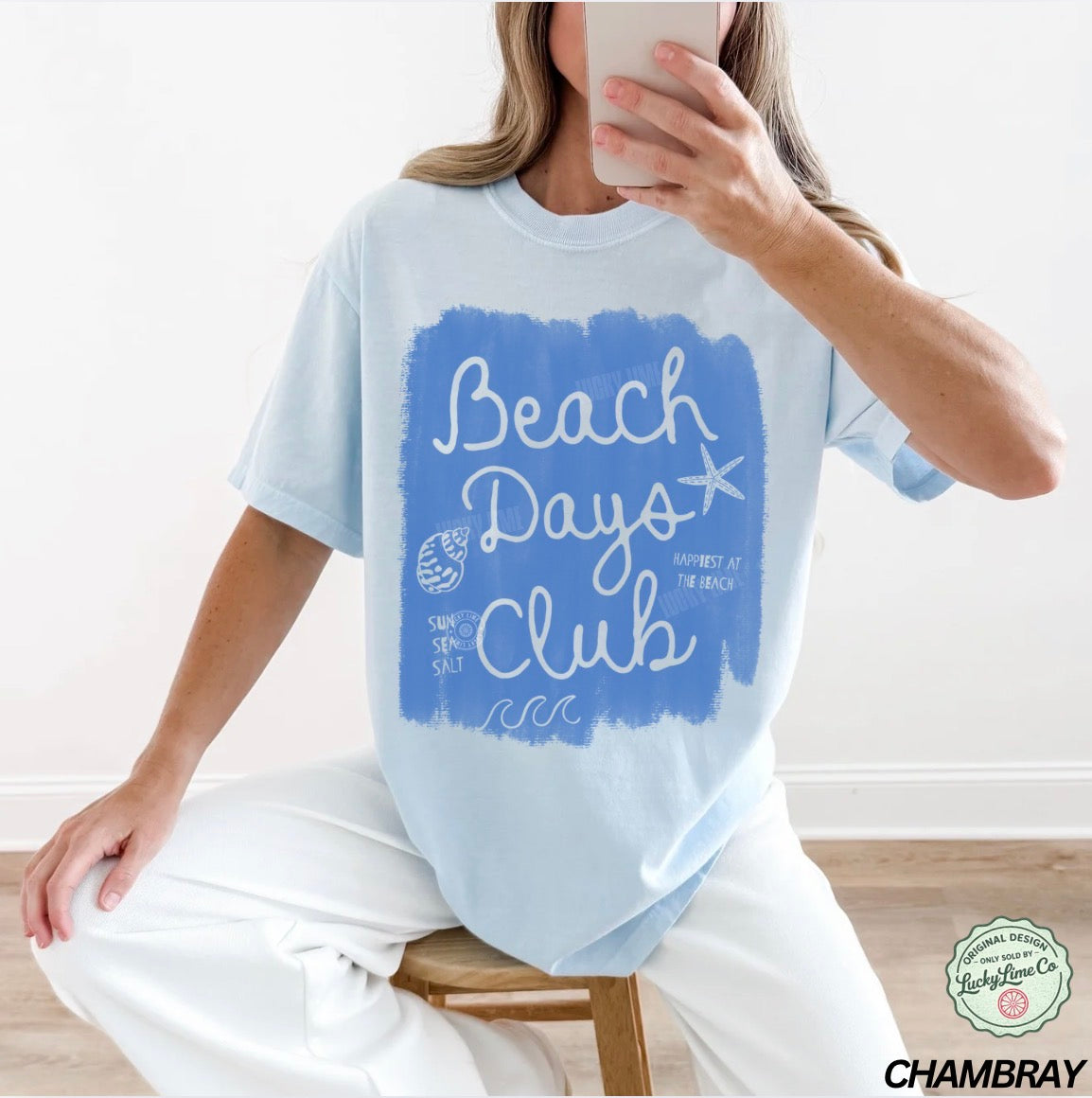 Person wearing a light blue t-shirt with 'Beach Days Club' design, sitting on a stool indoors.