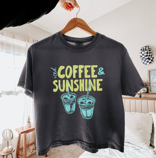 Black t-shirt with 'iced coffee & sunshine' text and graphics on a hanger in a room.