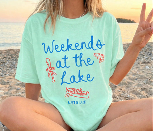 Person wearing a light blue t-shirt with 'Weekends at the Lake' text and graphics on a beach.
