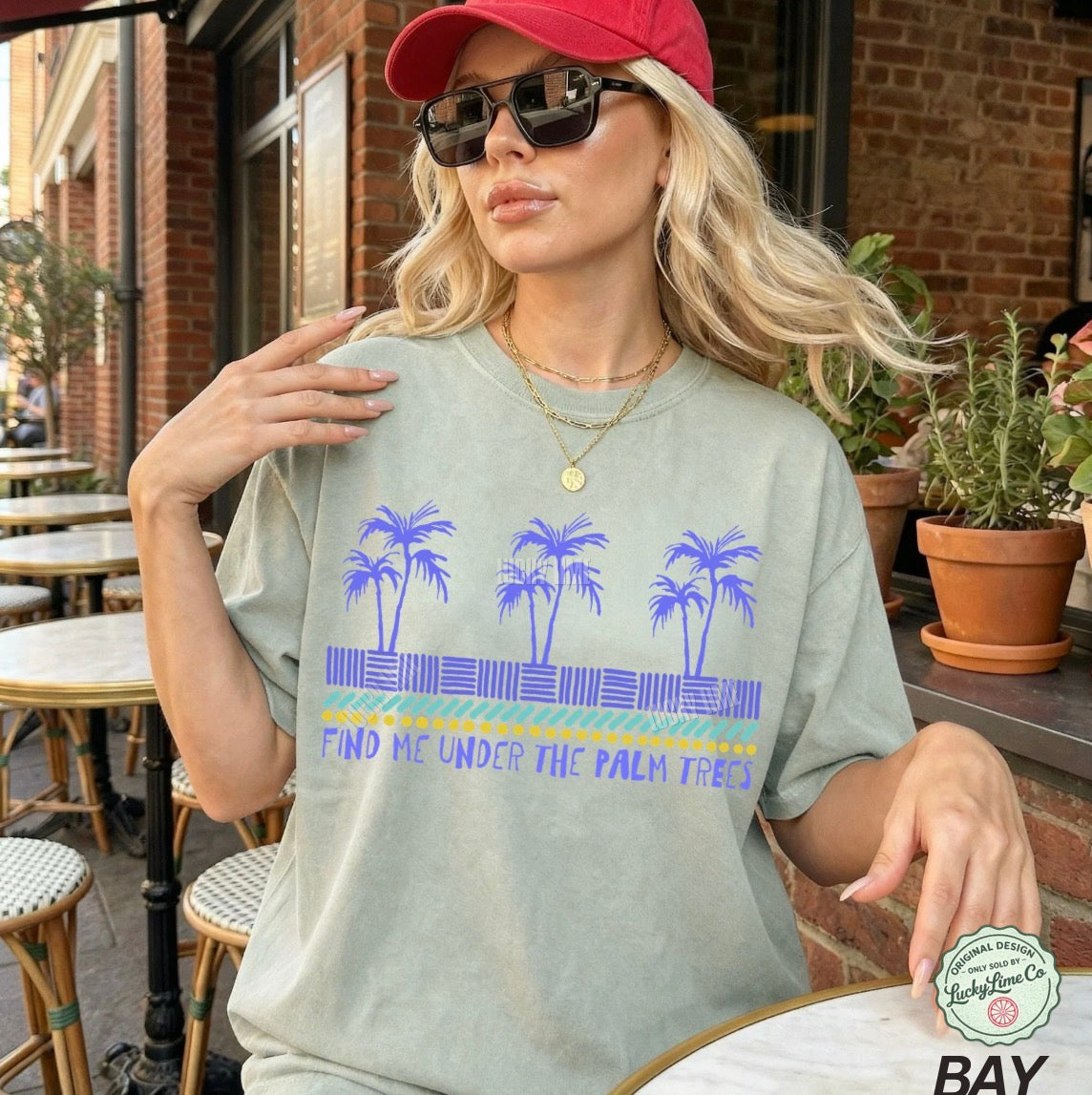 Woman wearing a t-shirt with palm tree design and text, sitting outdoors.
