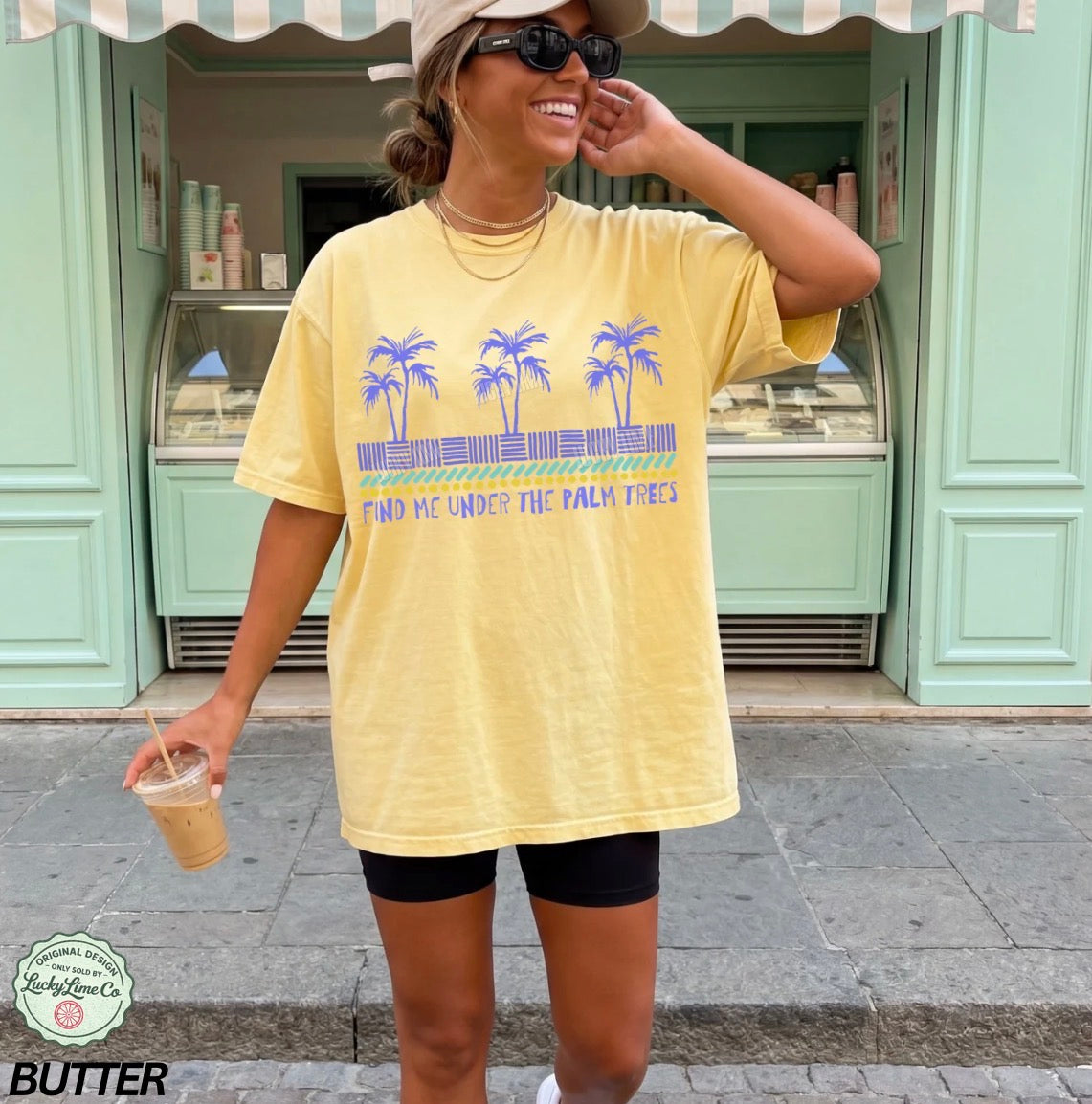Woman wearing a yellow t-shirt with palm tree design and text, standing in front of a green building.