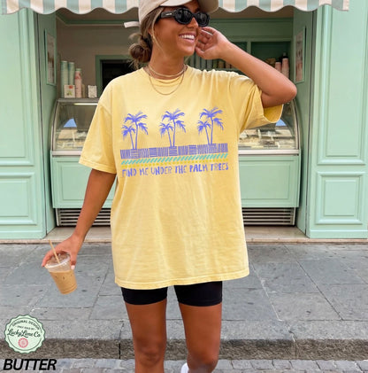 Woman wearing a yellow t-shirt with palm tree design and text, standing in front of a green building.