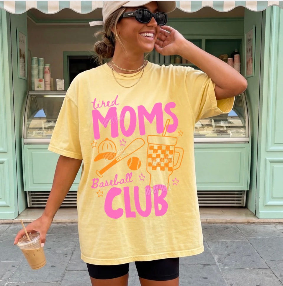 Woman wearing a yellow t-shirt with 'tired moms baseball club' text, standing in front of a light green building.