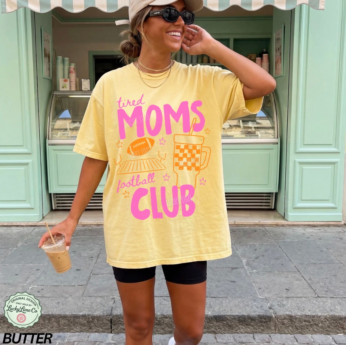Woman wearing a yellow t-shirt with 'tired moms football club' text, standing outside a store.