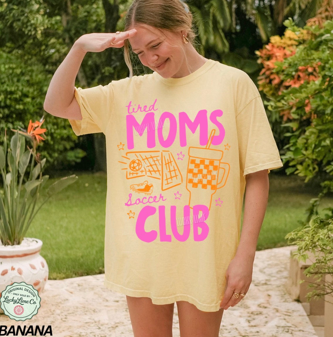 Woman wearing a yellow t-shirt with 'tired moms soccer club' text and graphics outdoors.