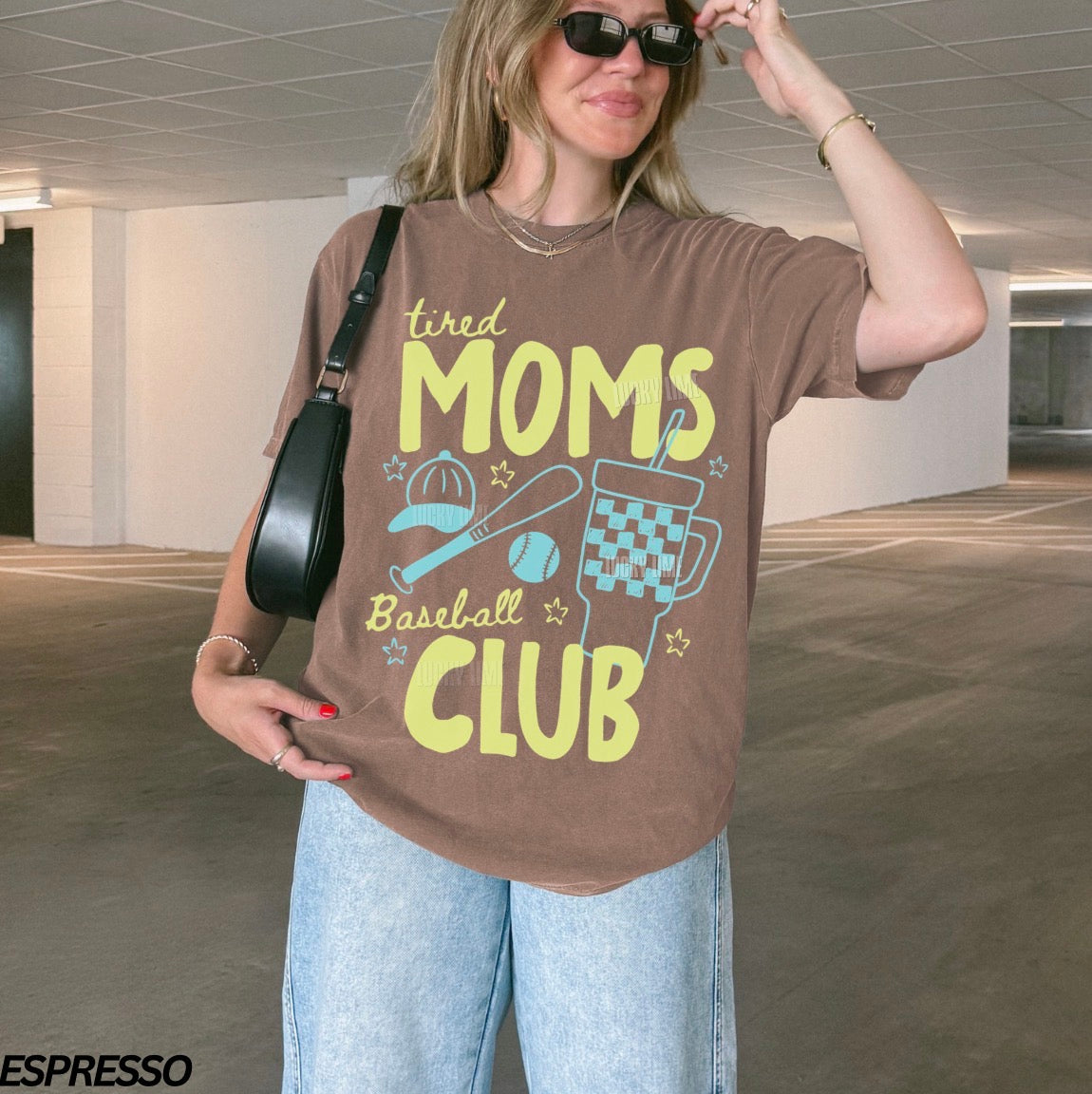 Woman wearing a brown t-shirt with 'tired moms baseball club' text in an indoor setting.