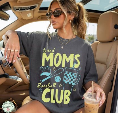 Woman wearing a 'tired moms baseball club' shirt in a car, holding a drink.