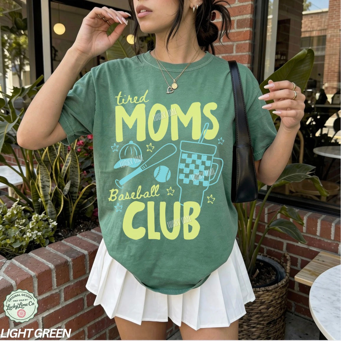 Person wearing a green t-shirt with 'tired moms baseball club' text and graphics outdoors.