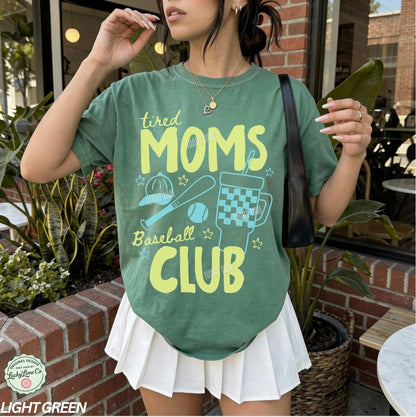 Person wearing a green t-shirt with 'tired moms baseball club' text and graphics outdoors.