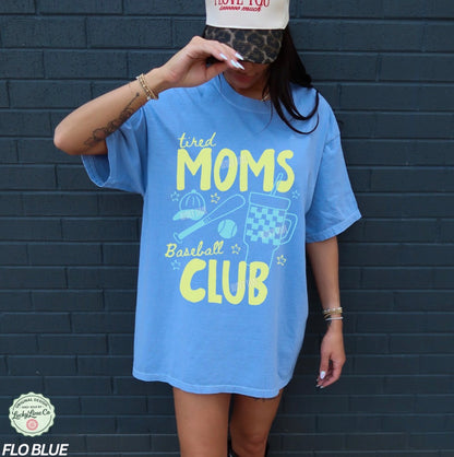 Person wearing a blue t-shirt with 'tired moms baseball club' text against a dark brick wall.