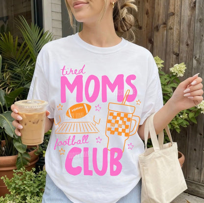 Person wearing a 'tired moms football club' shirt holding a drink and a bag outdoors.