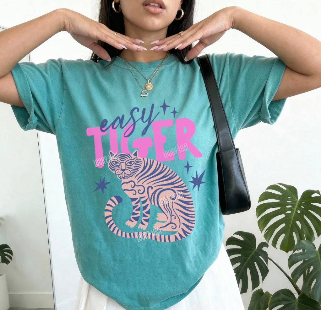 Person wearing a teal t-shirt with 'easy tiger' design, standing indoors with plants in the background.