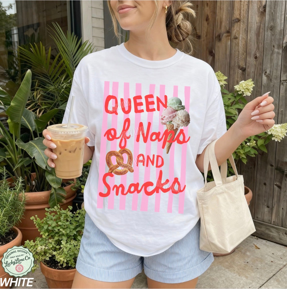 Person wearing a white t-shirt with 'Queen of Naps and Snacks' text and holding a drink outdoors.