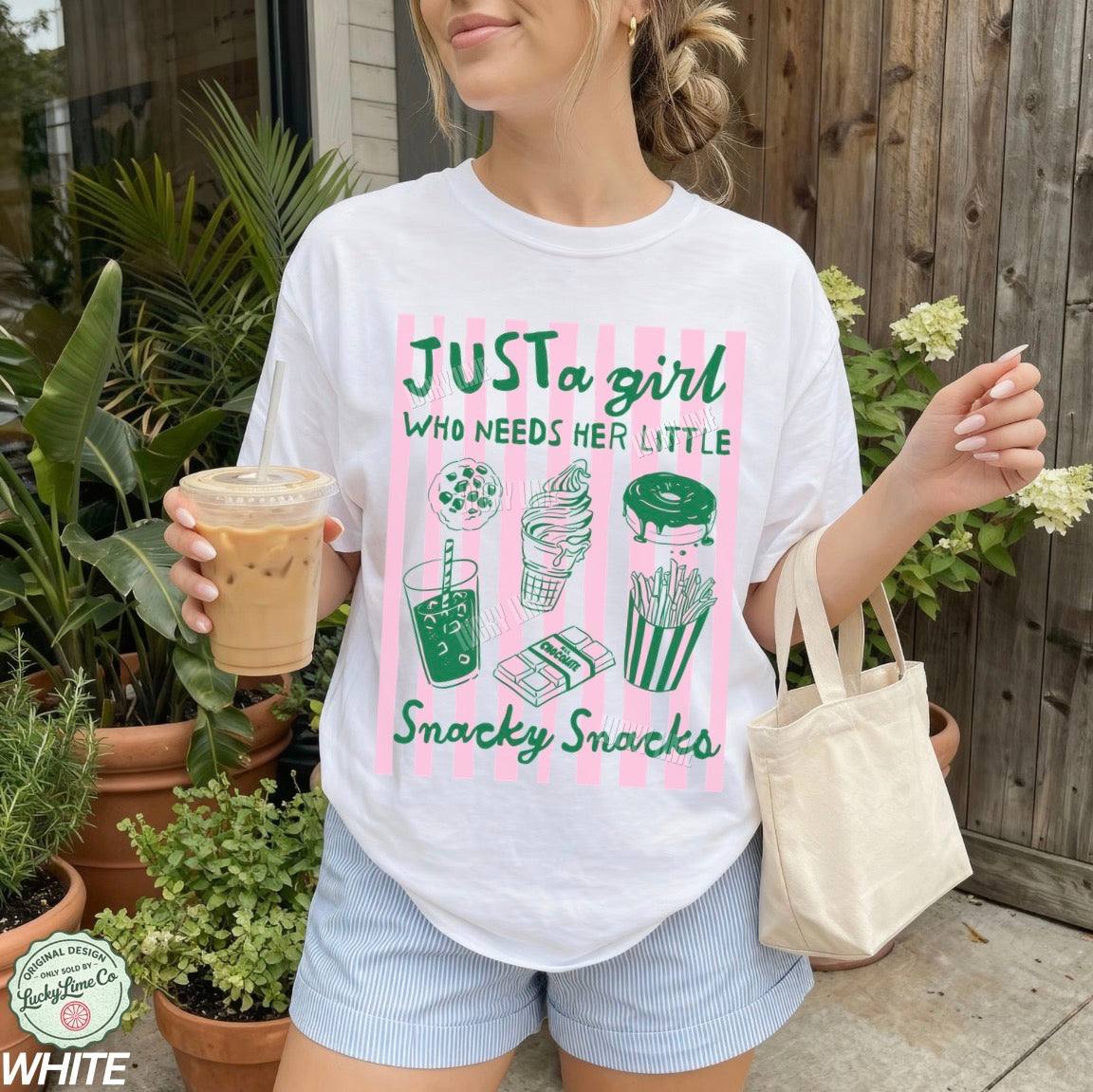 Woman wearing a white t-shirt with a graphic design holding a drink and a bag outdoors.