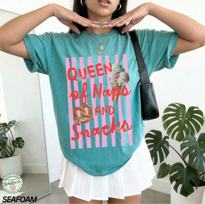 Person wearing a teal t-shirt with a colorful graphic and text, standing indoors with plants in the background.