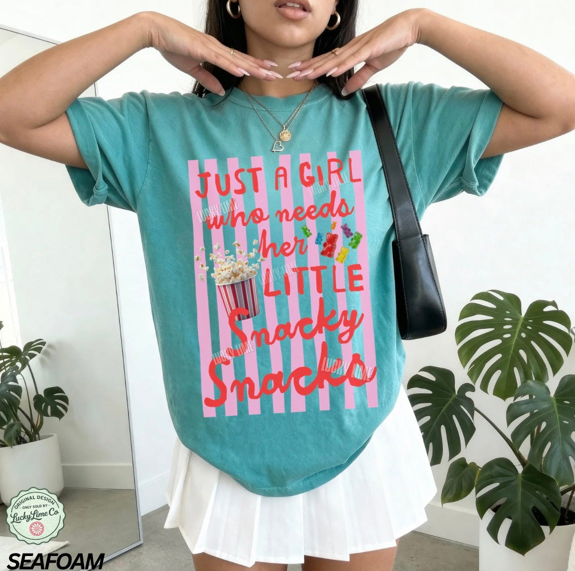 Person wearing a teal t-shirt with a colorful graphic and text, standing indoors with plants in the background.