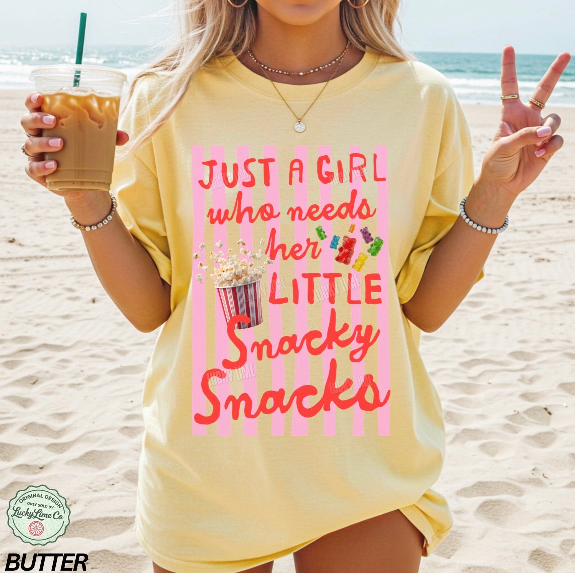 Woman on a beach wearing a yellow t-shirt with a snack-themed design, holding a drink.