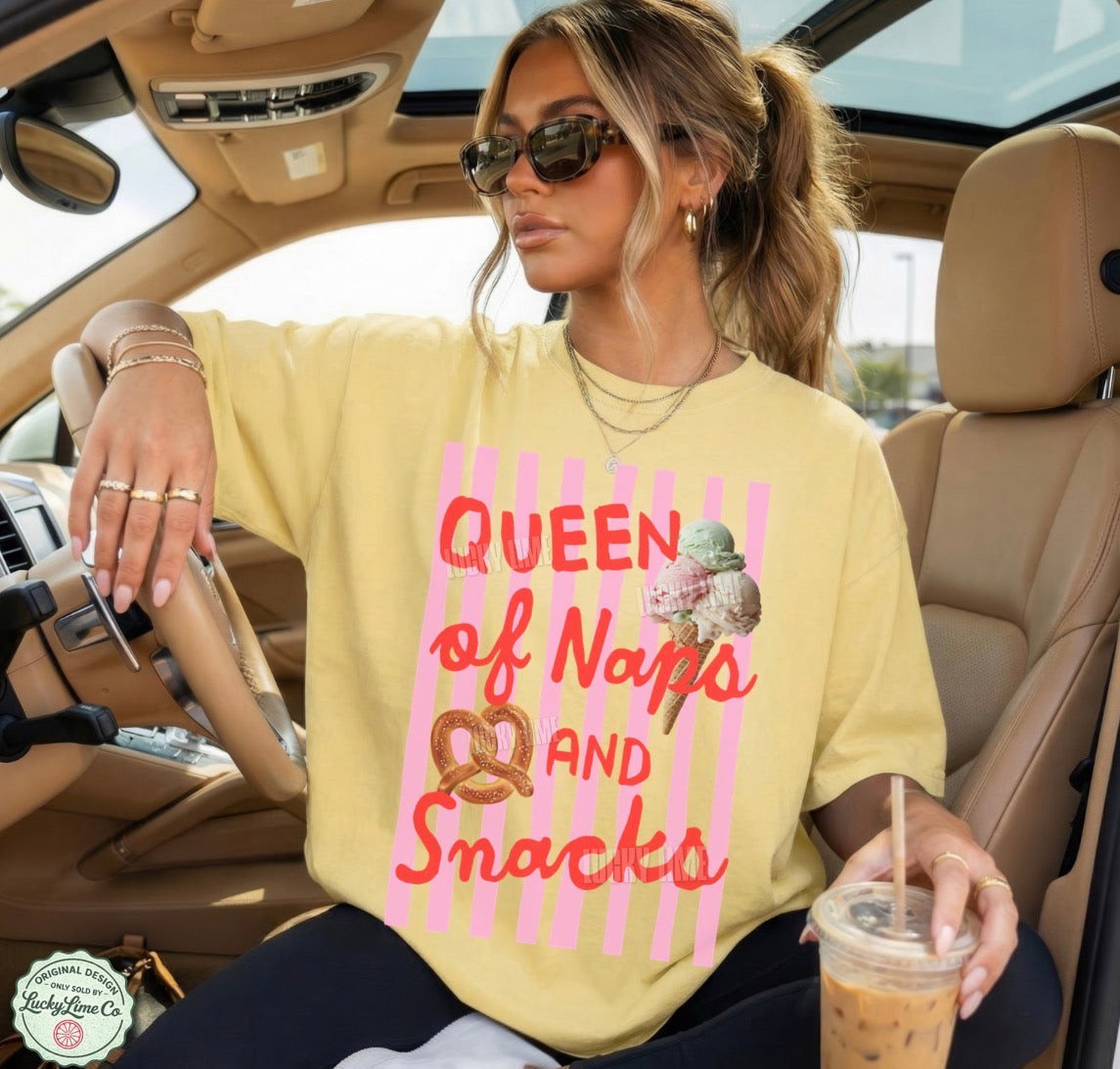 Woman sitting in a car wearing a yellow sweatshirt with text and ice cream graphics, holding a drink.