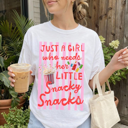 Woman wearing a t-shirt with a snack-themed design, holding a drink and a bag outdoors.
