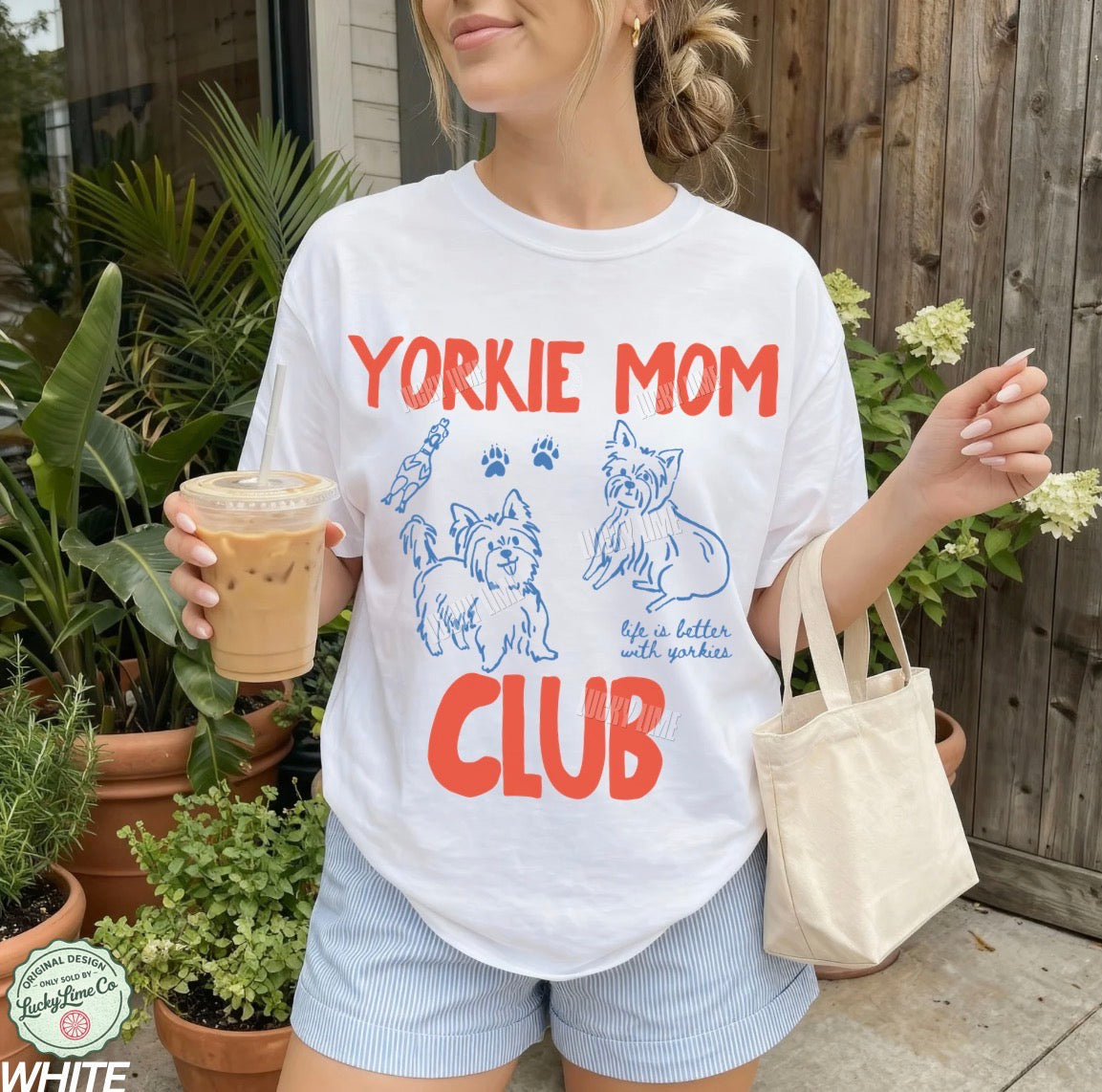 Person wearing a 'Yorkie Mom Club' t-shirt holding a drink and a bag outdoors.