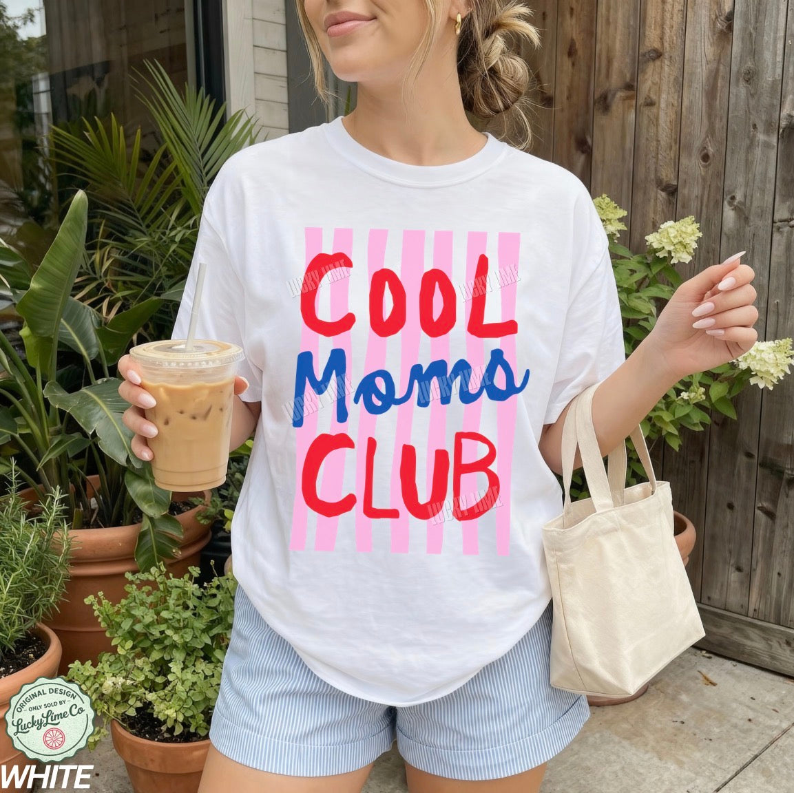 Person wearing a 'Cool Moms Club' t-shirt holding a drink and a bag outdoors.