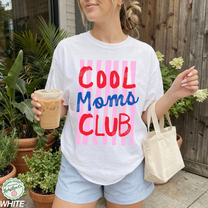 Person wearing a 'Cool Moms Club' t-shirt holding a drink and a bag outdoors.