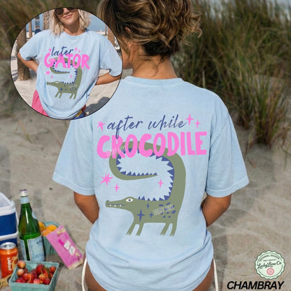 Person wearing a light blue t-shirt with a crocodile graphic and text on a beach.