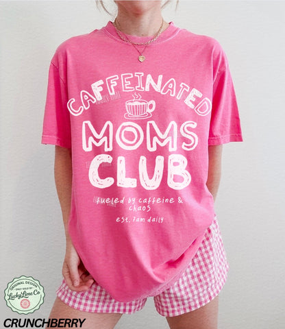 Person wearing a pink 'Caffeinated Moms Club' t-shirt and checkered shorts on a plain background