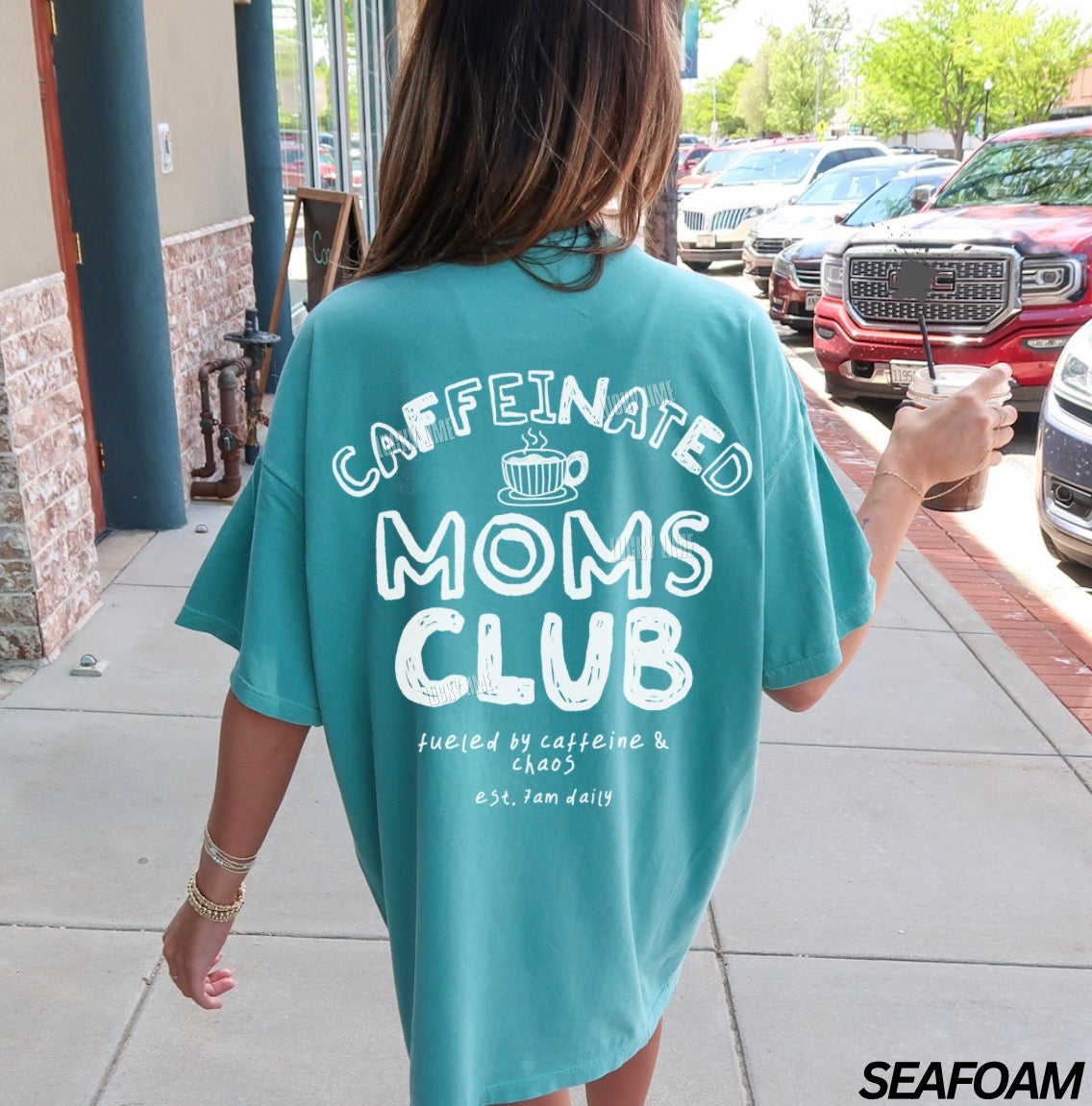 Person wearing a teal t-shirt with 'Caffeinated Moms Club' text, walking outdoors.