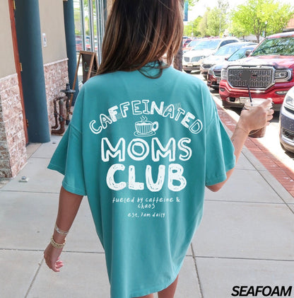 Person wearing a teal t-shirt with 'Caffeinated Moms Club' text, walking outdoors.
