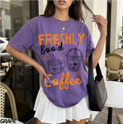 Person wearing a purple t-shirt with 'Freshly Bood Coffee' text and graphics outdoors.