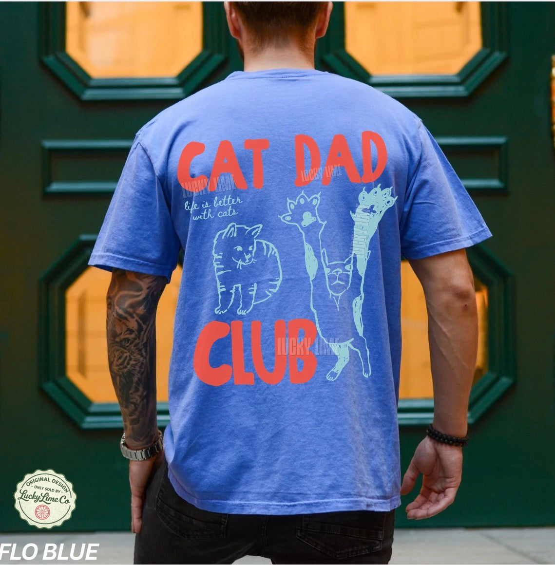 Person wearing a blue t-shirt with 'Cat Dad Club' text and graphics on the back, standing in front of a green door.