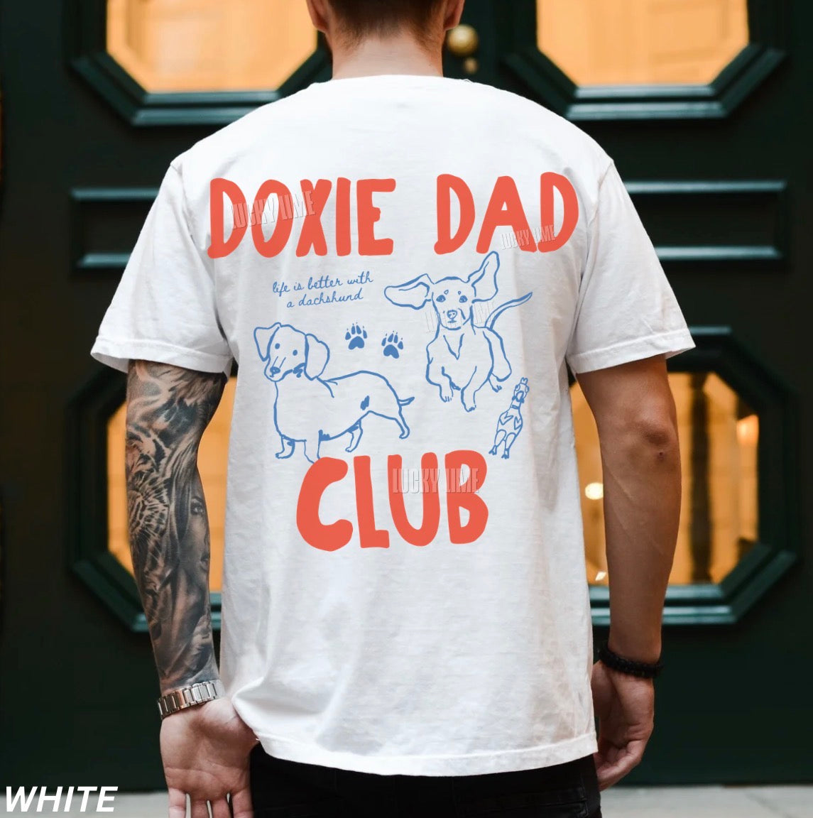 Person wearing a white t-shirt with 'Doxie Dad Club' design and tattoos on arm.