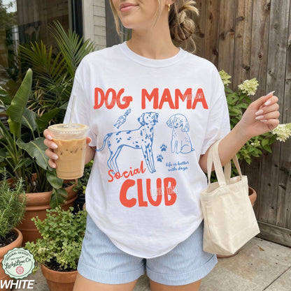 Person wearing a 'Dog Mama Social Club' t-shirt holding a drink and a bag outdoors.