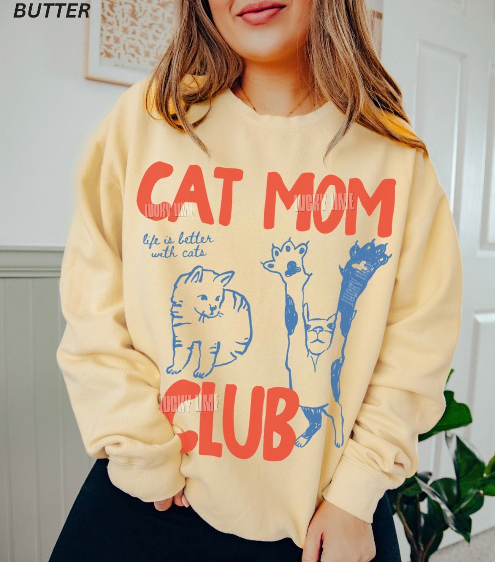 Beige hoodie with 'Cat Mom Club' text and cat illustrations worn by a person.
